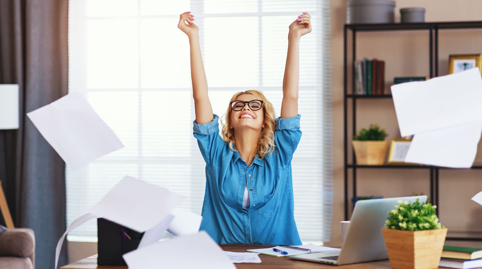 A blonde woman in a blue shirt throwing papers in the air cheerfully